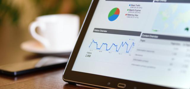 Tablet on a table with a coffee cup. On the tablet screen is several charts and graphs